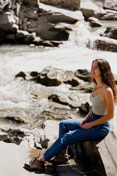 Traveler girl is sitting on the rock near waterfall and looking toward. Travelling in Karpathian mountains. Cascade waterfall. Beautiful landscape.