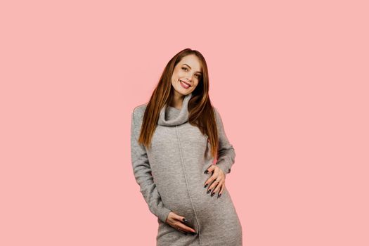 Pregnant woman. Expectation of the child. Maternity leave. Pregnancy of a happy beautiful girl in a gray sweater on a pink background