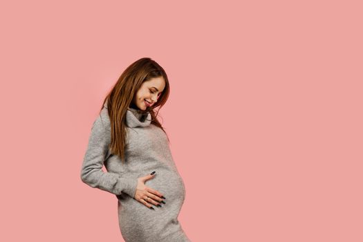 Pregnant woman. Expectation of the child. Maternity leave. Pregnancy of a happy beautiful girl in a gray sweater on a pink background