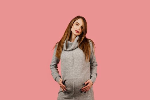 Pregnancy of a happy beautiful girl in a gray sweater on a pink background. Pregnant woman. Expectation of the child. Maternity leave.