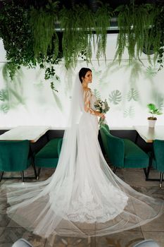 Young attractive bride in wedding dress with bridal veil looks at bouquet and smiles. Girl in wedding gown in modern restaurant.