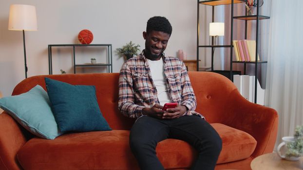 Cheerful young man sitting on sofa, using mobile phone share messages on social media application