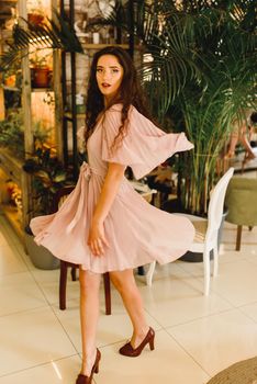Young woman in soft pink dress is moving and looking into the camera. Beauty girl in cafe with big green plants.