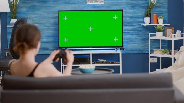 Young woman holding wireless controller playing action console video game on green screen tv