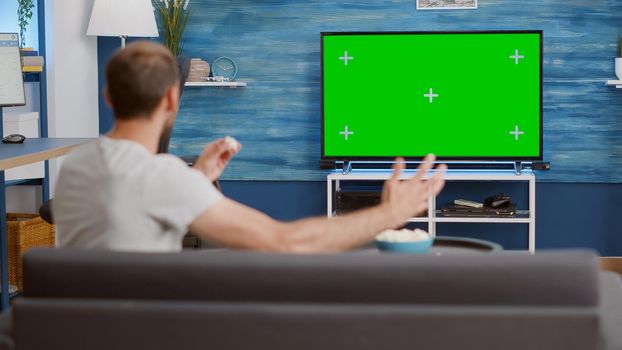 Sport fan sitting on sofa watching sport game on green screen tv mockup encouraging favourite team