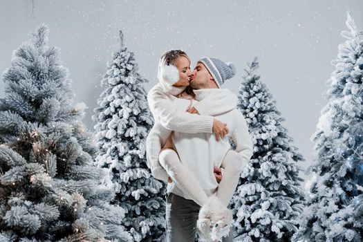 Couple kiss and hug. Man holds girl near christmas trees in winter day. New year celebration. People weared wearing fur headphones, hats, white sweaters
