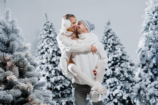 Couple couple laughing and having fun while snow falls near christmass trees. Winter holidays. Love story of young couple weared white pullovers. Happy man and young woman hug each other