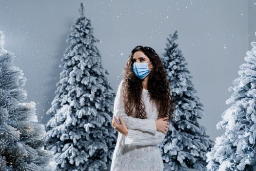 New year celebration at covid-19 coronavirus quarantine period. Happy girl in medical mask with falling snow stays at home. Social distance. Winter holidays in snowy day.