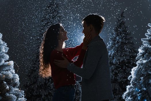 Falling snow and kisses with falling snow on dark blue background. Happy young couple hug and kiss near christmas trees at the eve of new year celebration in winter day.