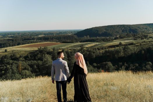 Muslim love story. Mixed couple smiles and hugs on the green hills . Woman weared in hijab looks to her man. Advert for on-line dating agency.