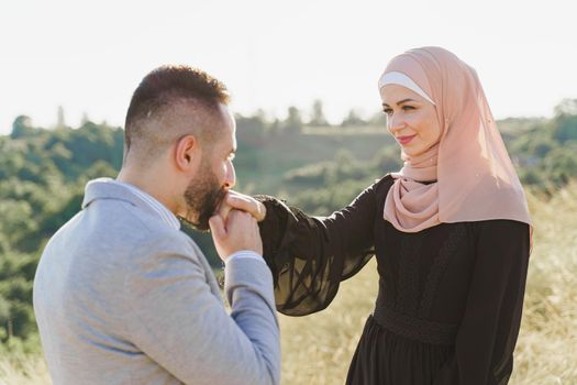 Muslim love story close-up. Mixed couple smiles and hugs on the green hills . Woman weared in hijab looks to her man. Advert for on-line dating agency.