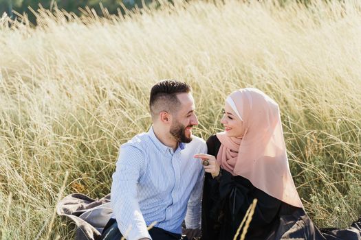 Muslim love story. Mixed couple seats on the grass, smiles and hugs.. Woman weared in hijab looks to her man. Advert for on-line dating agency.