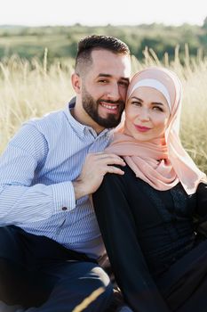 Muslim love story close-up. Mixed couple smiles and hugs on the green hills . Woman weared in hijab looks to her man. Advert for on-line dating agency.
