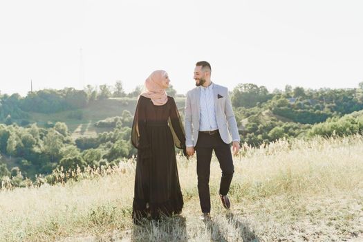 Muslim love story of mixed couple. Man and woman smiles and walks on the green hills . Woman weared in hijab looks to her man. Advert for on-line dating agency.