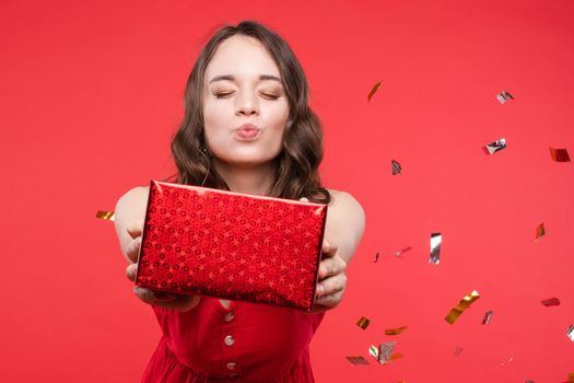 Pleasant young woman taking gift box kissing with closed eyes at red studio background
