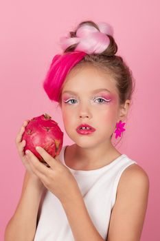 Close-up portrait of pretty girl with pink hairstyle with dragon fruit on pink background. Studio shot of charming tween girl with pink make up enjoying juicy red pitaya. exotic Pitahaya fruit