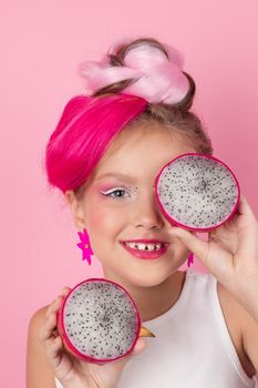 Close-up portrait of pretty girl with pink hairstyle with dragon fruit on pink background. Studio shot of charming tween girl with pink make up enjoying juicy red pitaya. exotic Pitahaya fruit