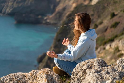 Woman tourist enjoying the sunset over the sea mountain landscape. Sits outdoors on a rock above the sea. She is wearing jeans and a blue hoodie. Healthy lifestyle, harmony and meditation
