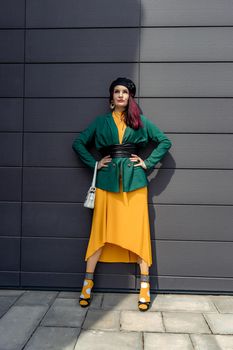 Young woman in bright clothes, yellow skirt and green jacket. Yellow socks in sandals, beret on the head, hair with the color of magenta. Caucasian female fashion model standing against gray wall background, open with copy space.