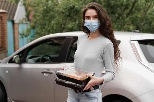 Sushi set in box healthy food delivery service by car. Girl courier in medical mask with 2 sushi boxes stands in front of car.