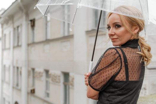 The blonde stands under a transparent umbrella during the rain. The fall season. Rear view. The woman is dressed in a black lace dress, her hair pulled back in a ponytail.