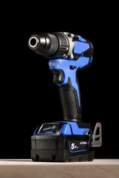 Cordless drill stands on a wooden table on a black background. Cordless drill with lithium-ion battery in blue. Professional tool for drilling holes and driving screws.