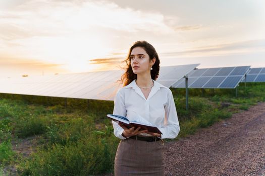 Investor woman stand and read book near solar panels row on the ground at sunset. Sustainability of planet.