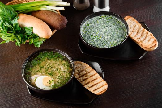 Okroshka, broth with noodles and eggs in black plate, toasted bread, herbs on dark wooden table. Healthy seasonal lunch.