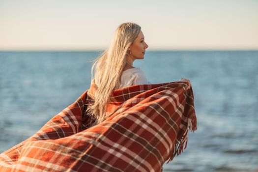 Attractive blonde Caucasian woman enjoying time on the beach at sunset, walking in a blanket and looking to the side, with the sunset sky and sea in the background. Beach vacation