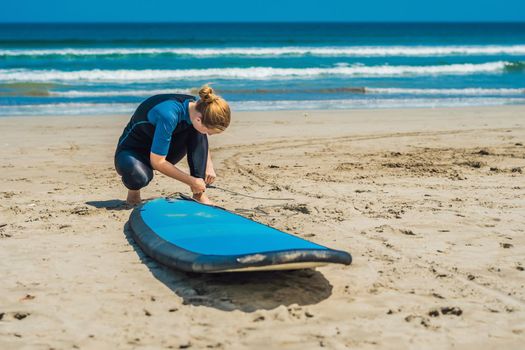 Summer time and active rest concept. Young surfer woman beginner fastens leash across leg, going to surf on big barral waves on ocean, dresseed in boardies, poses aganist sky on hot sunny day