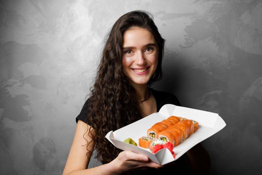 Girl holding philadelphia rolls in a paper box on gray background. Sushi, food delivery.
