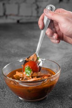 Soup with meat, mushrooms, tomatoes in transparent bowl on gray background. Appetizing thick satisfying tom yam.