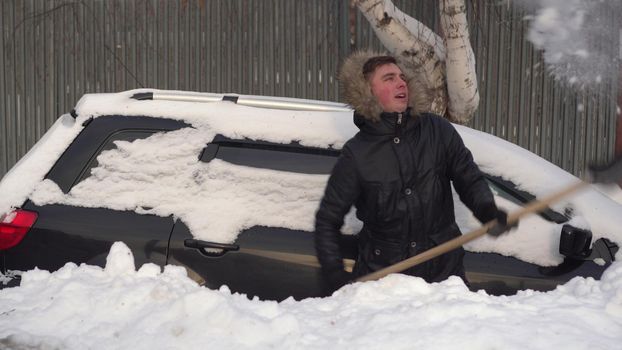 A young man in a jacket digs his car out of a snowdrift with a shovel. The car was covered in snow. Weather disaster.