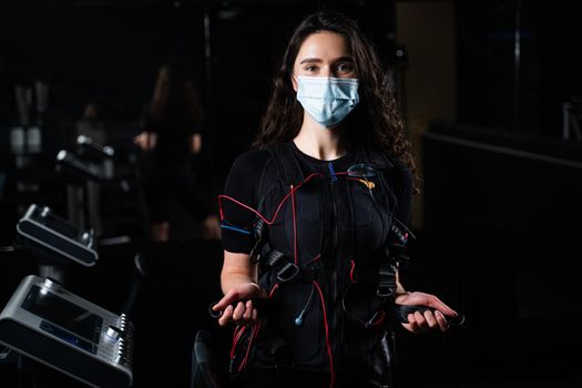 Girl in EMS suit and medical mask in gym. Protection from coronavirus covid-19. Sport training in electrical muscle stimulation suit at quarantine period.