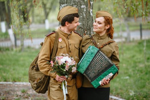 People in Soviet military uniforms. A couple stands by a tree, a soldier embraces a military girl playing an accordion