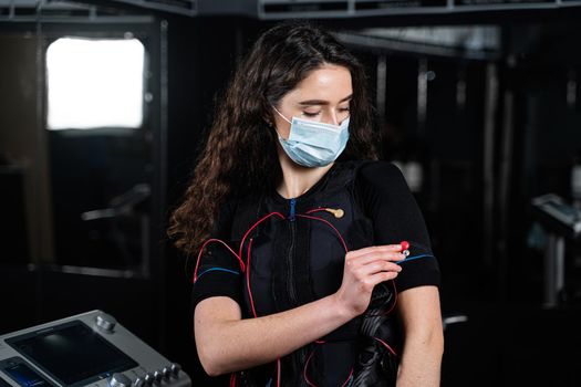 Girl in EMS suit and medical mask in gym. Protection from coronavirus covid-19. Sport training in electrical muscle stimulation suit at quarantine period.