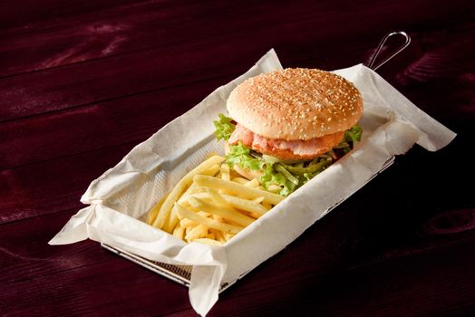 Burger with cutlet, bacon, salad, pickled cucumber and potato fries on parchment in a metal basket on a wooden background