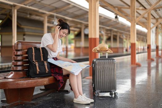 young asian woman traveler sitting with map choose where to travel and bag waiting for train at train station, summer vacation travel concept