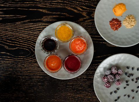 Assortment of sauces, spices, frozen berries on gray plates on a dark wooden table. View from above