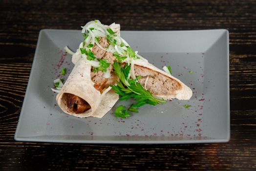 Lula kebab roll in pita bread garnished with onions and cilantro leaves on a gray square plate on a dark wooden table. Georgian dish