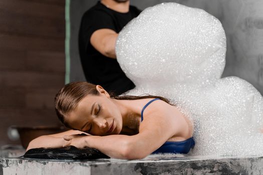 Attractive girl relaxing in spa. Masseur is making foam peeling in Turkish spa hammam.