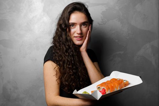Surprised girl with sushi set philadelphia rolls in a paper box happy girl holding on a gray background. Food delivery.