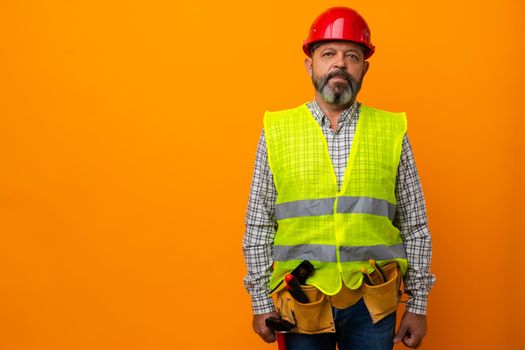 Middle-aged bearded man builder in uniform and hardhat with tools against orange background