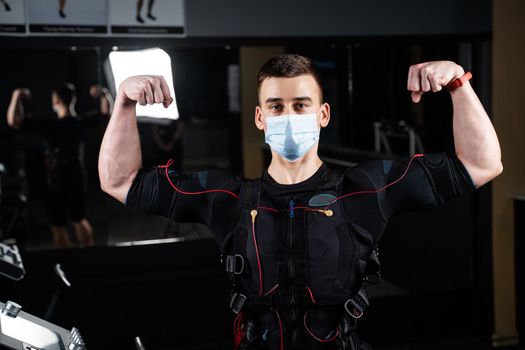 Man in EMS suit and medical mask in gym. Protection from coronavirus covid-19. Sport training in electrical muscle stimulation suit at quarantine period.