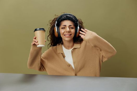 COOL MOOD CONCEPT. Enjoyed tanned curly Latin female in headphones with coffee cup to-go dance sing sit at table isolated over olive green background. Copy space Mockup Banner. Fashion offer
