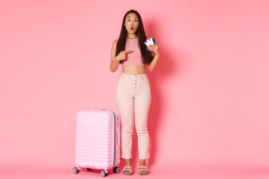 Travelling, holidays and vacation concept. Full-length of surprised and amazed girl tourist standing near suitcase, pointing finger at airline flight tickets and passport, pink background