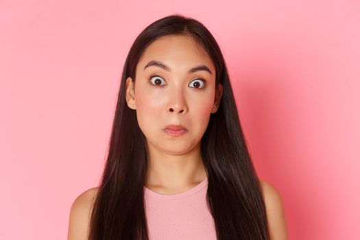 Beauty, fashion and lifestyle concept. Close-up of ambushed and startled asian brunette girl looking stupified, pulling face and raise eyebrows astounded, standing awkward over pink background