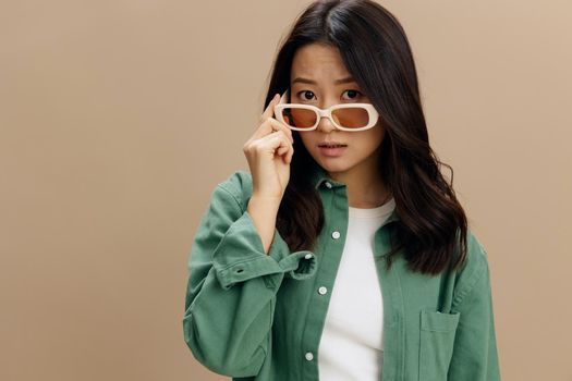 Closeup portrait of adorable Korean young woman in khaki green shirt touch stylish eyewear looks at camera posing isolated on beige pastel studio background. Cool fashion offer. Sunglasses ad concept