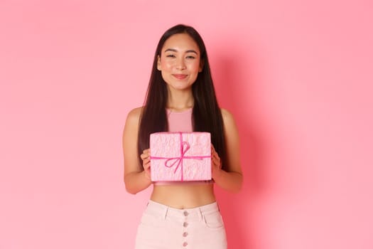Holidays, celebration and lifestyle concept. Close-up of pleased cheerful asian girl receive cute wrapped gift and smiling satisfied, likes receiving presents, standing over pink background.
