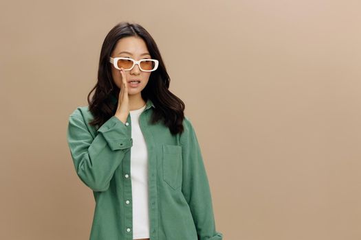 Shhh. Excited shocked Korean young woman in khaki green shirt stylish eyewear tells gossip aside posing isolated on over beige pastel studio background. Cool fashion offer. Sunglasses ad concept